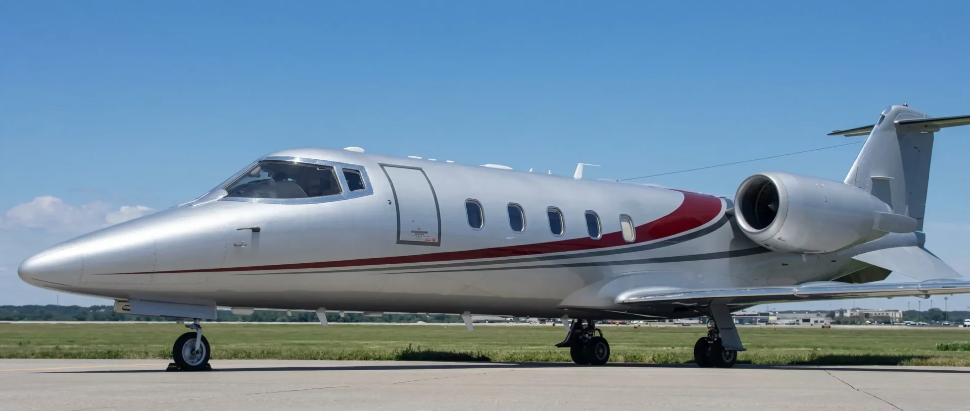Learjet 60 private jet with white and silver fuselage, red accent stripe, parked on airport tarmac under clear blue sky